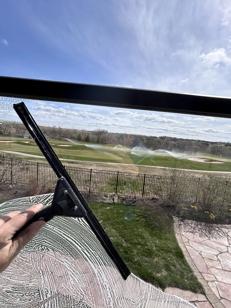 Squeegee pulling water off a deck railing glass panel with a clear view of a green backyard